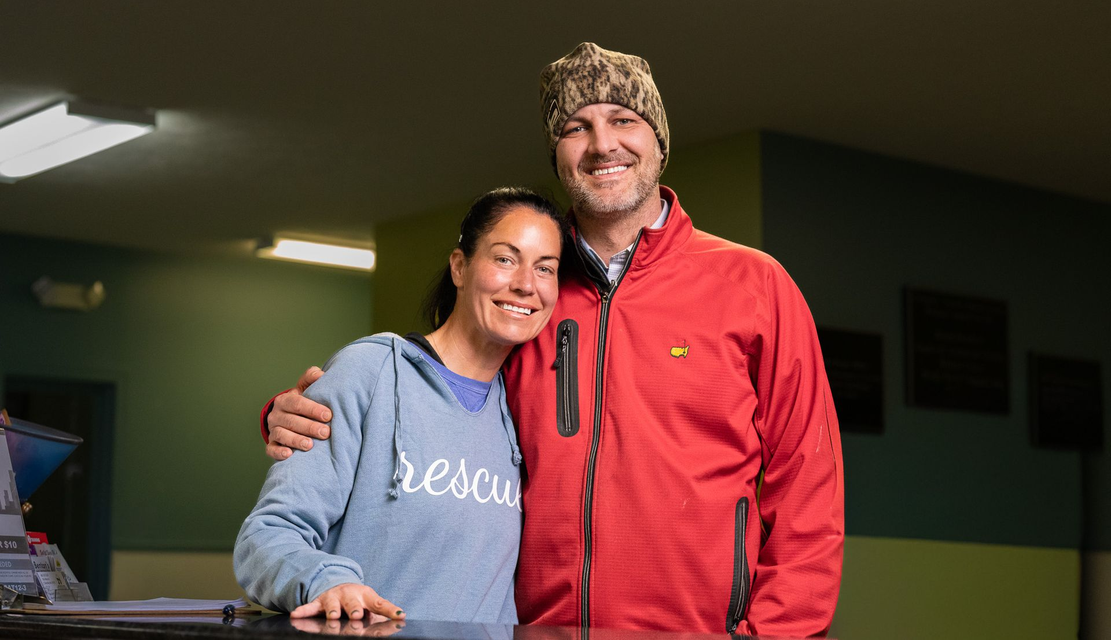 A smiling couple poses indoors, with the person on the left wearing a blue "rescue" hoodie and the other in a red jacket.
