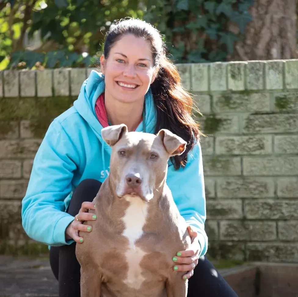 A smiling person wearing a blue hoodie sitting outdoors with a tan and white dog in front of a brick wall.