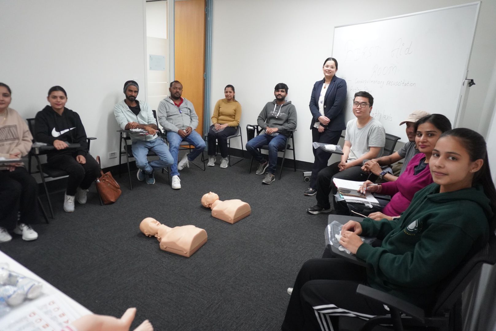 People in a classroom setting, participating in a CPR training session. Two mannequins are on the floor.