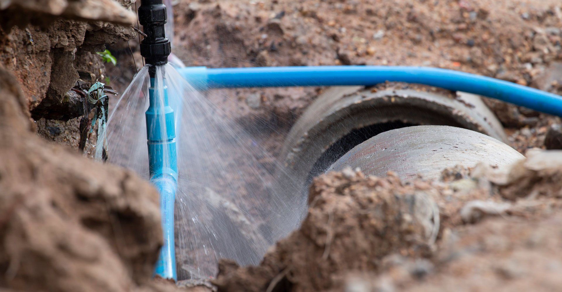 Water gushing from a broken blue pipe in a dirt trench, near a concrete pipe.