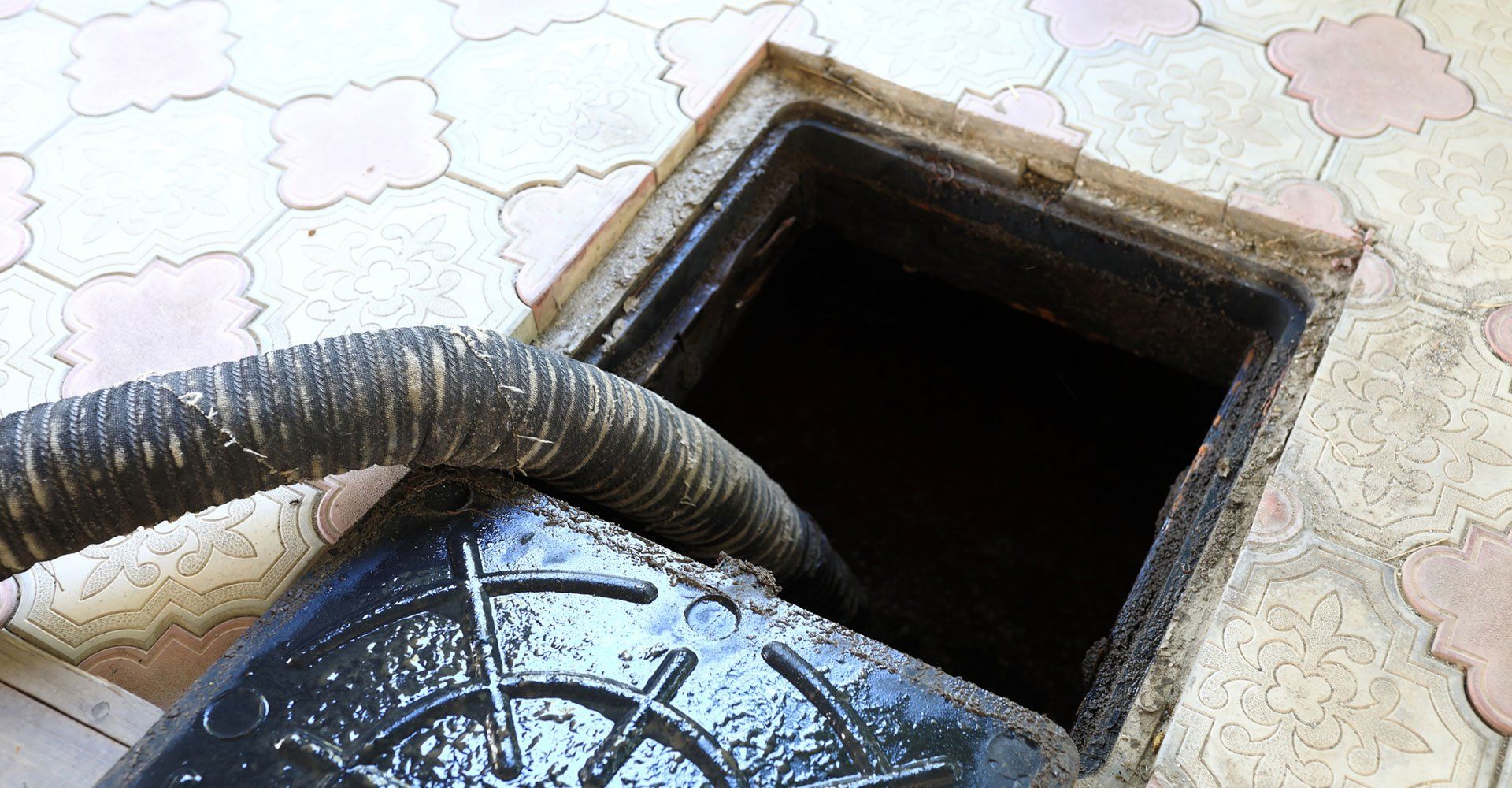 A hose pumping sludge from an open, square manhole cover on a patterned tile surface.