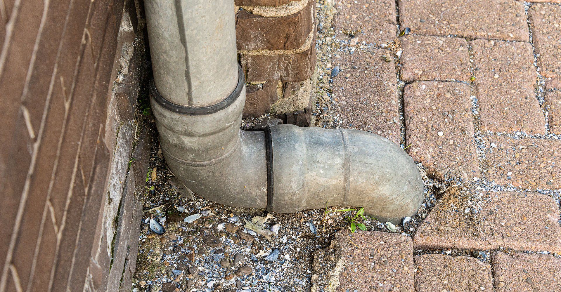 Gray downspout bends near a brick building and pavement; small debris is scattered around the base.