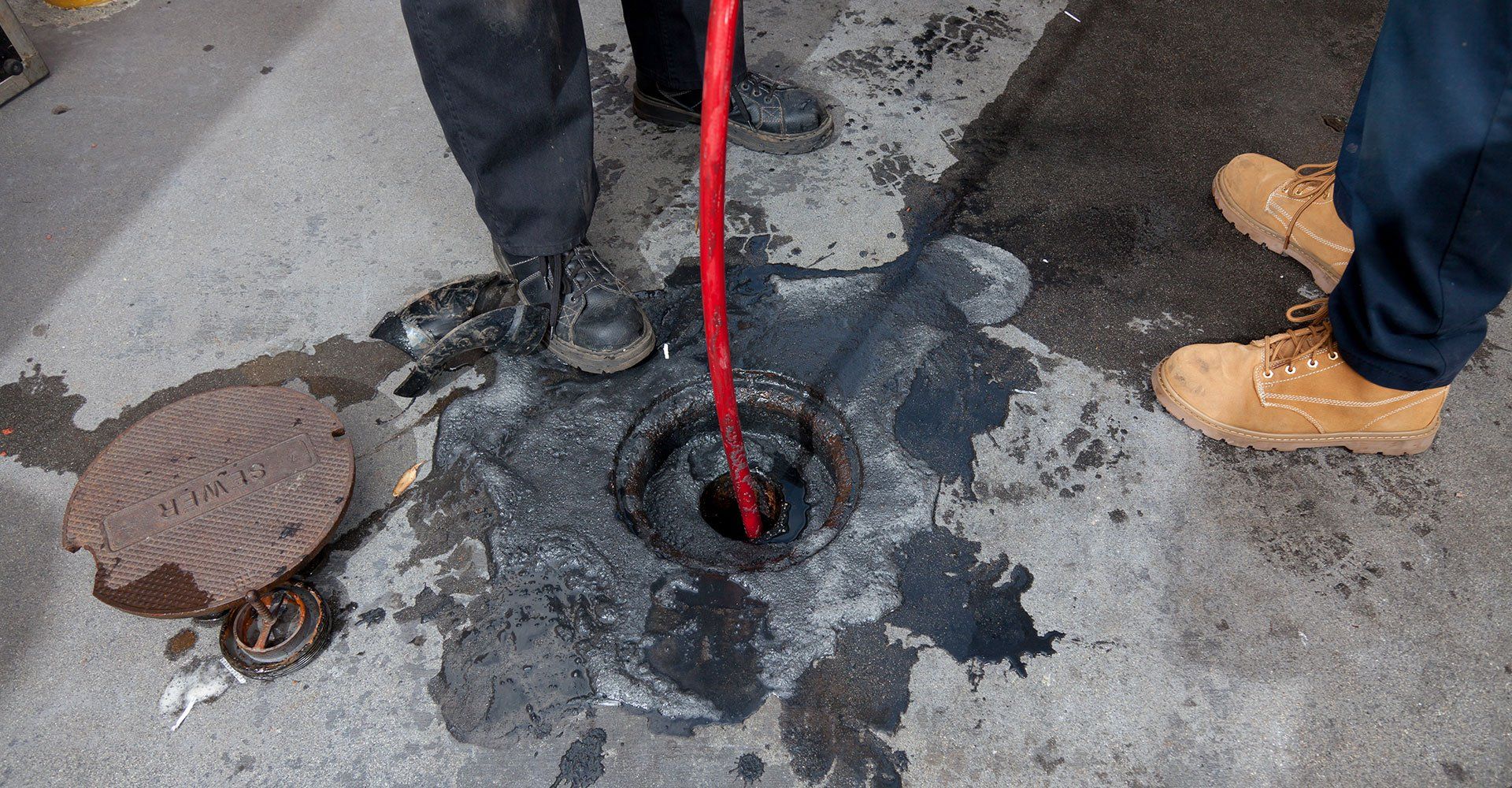 Workers with a red tool clearing a clogged drain in a concrete setting.