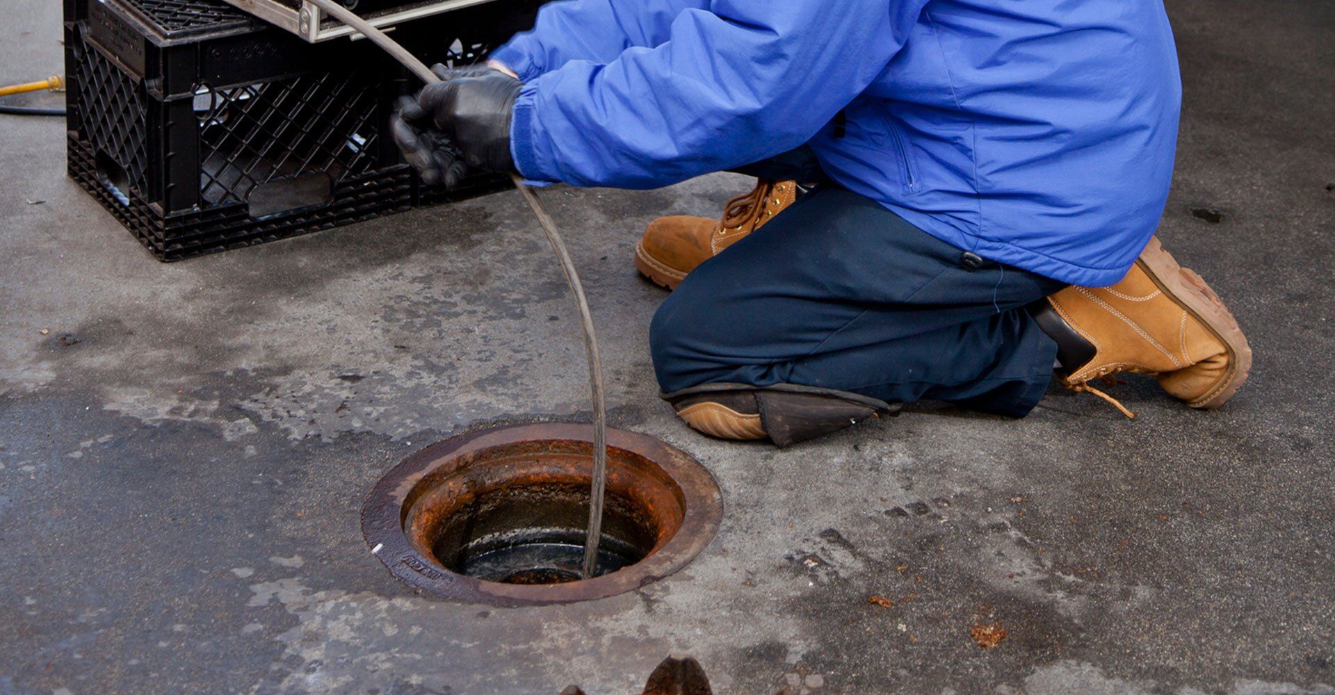 Person in blue jacket kneels, using a drain snake on a manhole in a concrete setting.