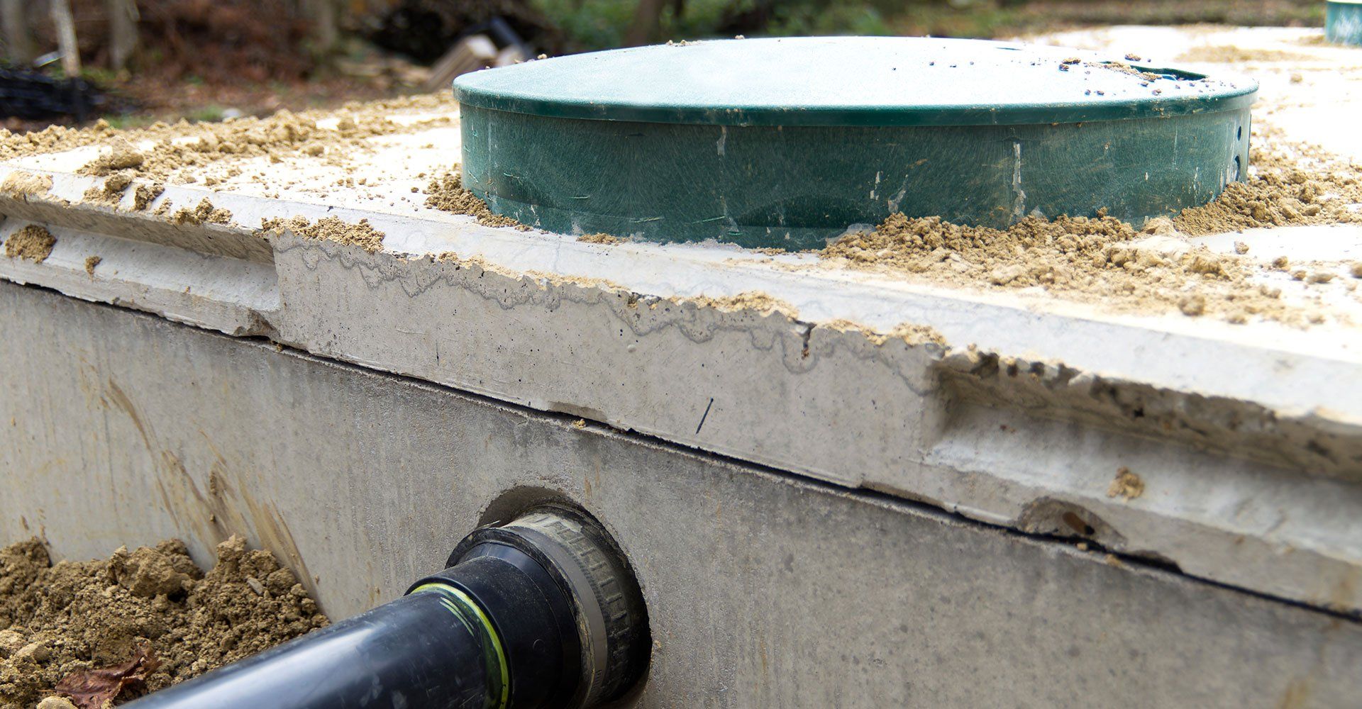 Close-up of a concrete septic tank with a green lid, black pipe, and dirt.