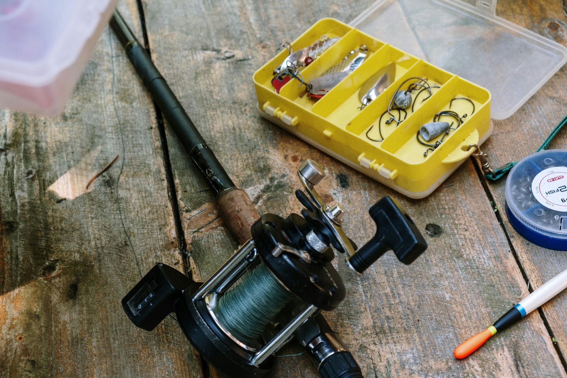 Fishing reel, tackle box, line spool, and pen on a weathered wooden surface.