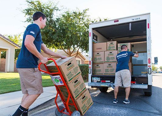Two movers loading boxes onto a truck using a hand truck on a sunny residential street.