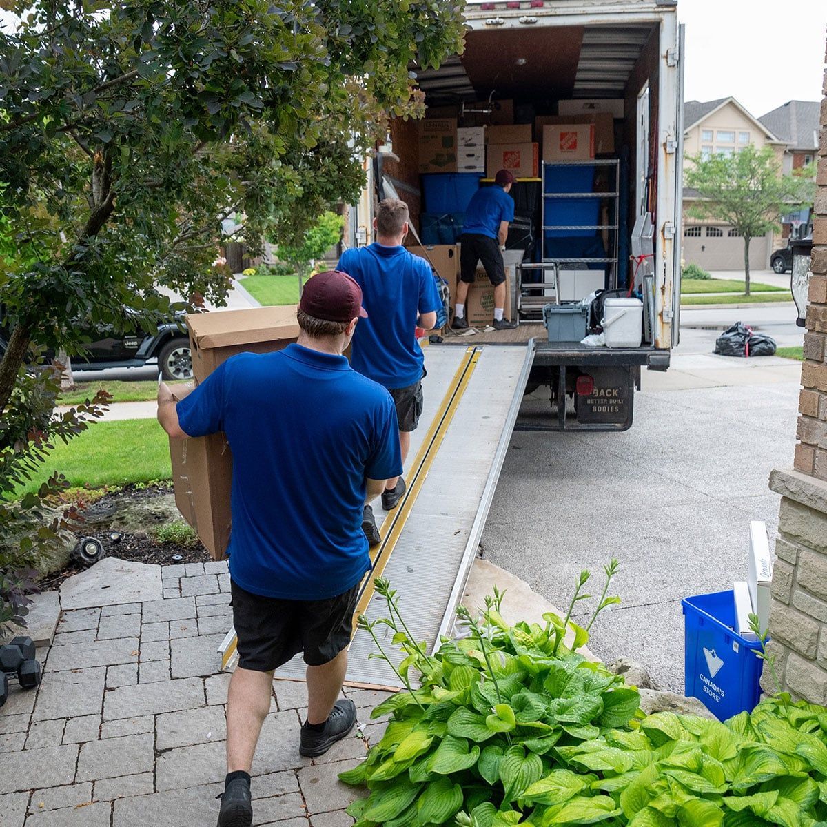 Movers carrying boxes to a truck parked in front of a house. Men in blue shirts and black shorts.