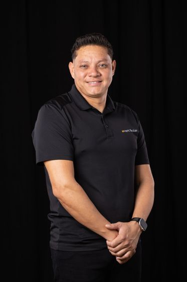 Man in a black polo shirt smiling against a dark background, hands clasped in front.