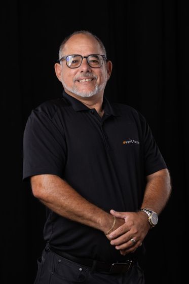 Man in black polo shirt and glasses standing against a black background, smiling with hands clasped.
