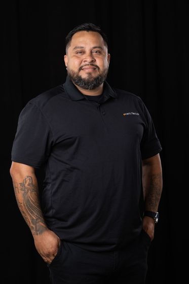 Portrait of a man in a black polo shirt standing against a dark background.