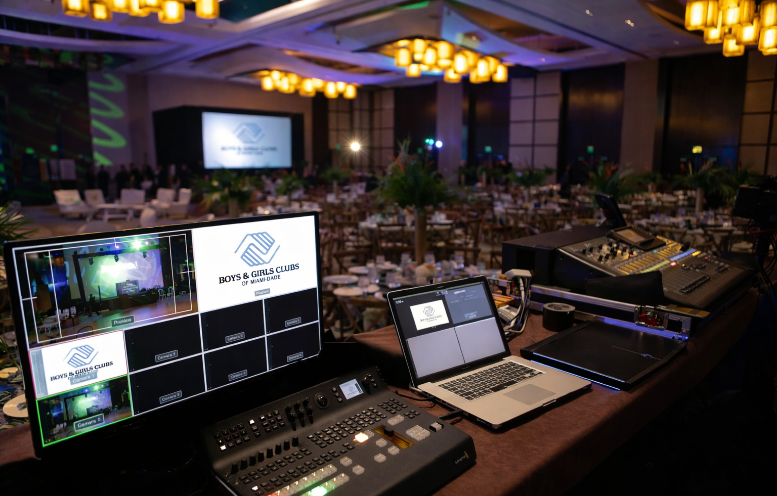 AV control booth overlooking a lit conference hall, with monitors, laptop, and audio/video equipment set up.