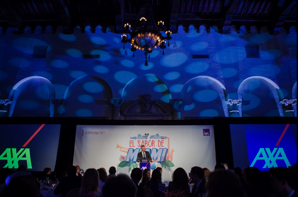 Conference stage with blue lighting, chandelier overhead, and a large screen flanked by AXA logos.