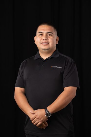 Person in a black polo shirt standing against a dark background with hands clasped.