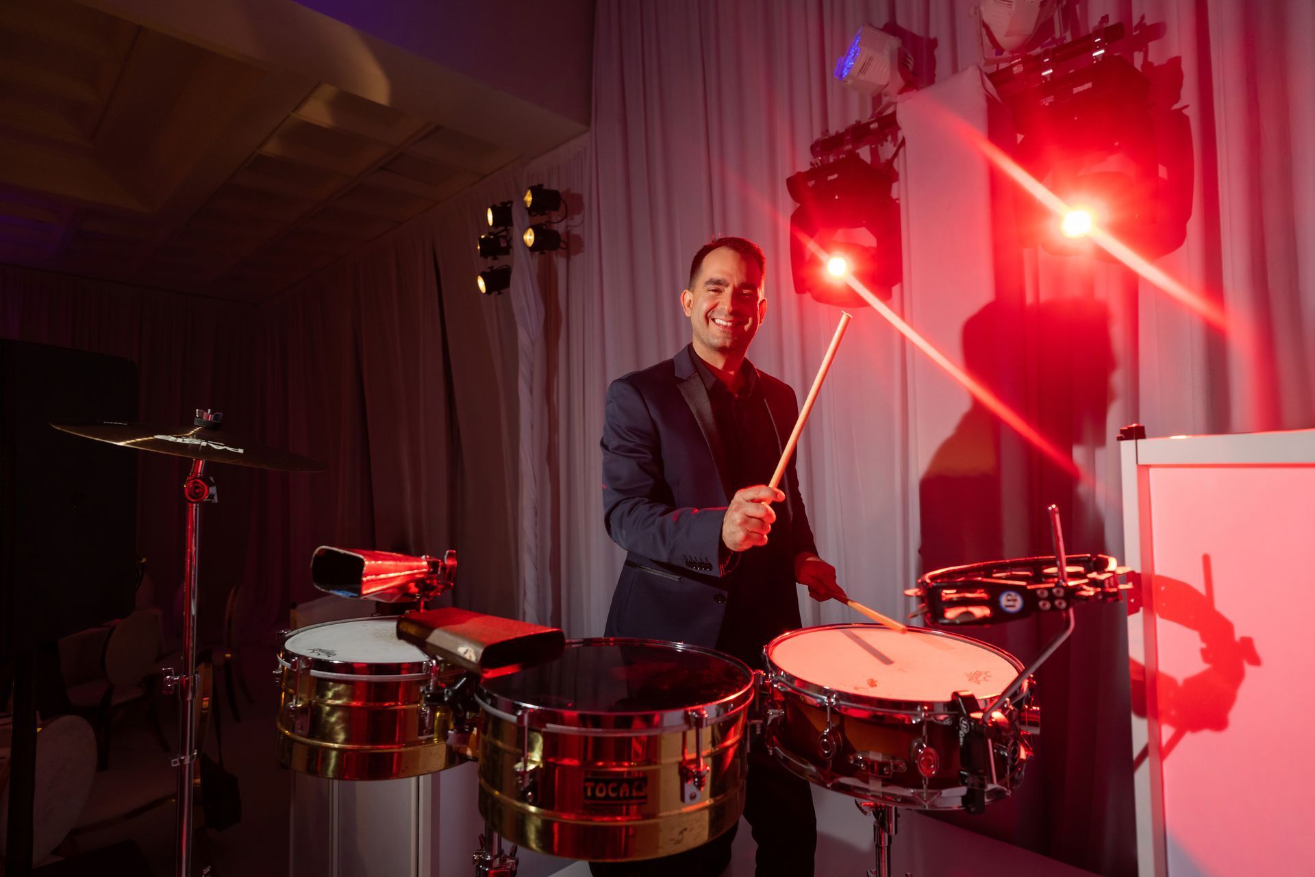 Smiling drummer playing timbales on a colorful lit stage with purple and orange lights