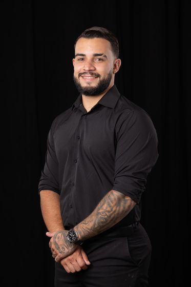 Man in a black shirt posing against a dark background, with tattooed forearms and hands clasped in front.