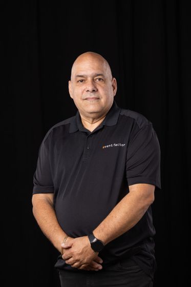 Man standing against a black background, wearing a black polo shirt and holding his hands together.