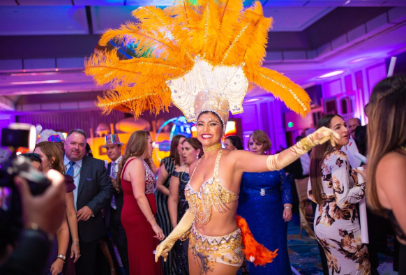 Dancer in a bright orange feathered costume performing at a lively indoor party with guests watching