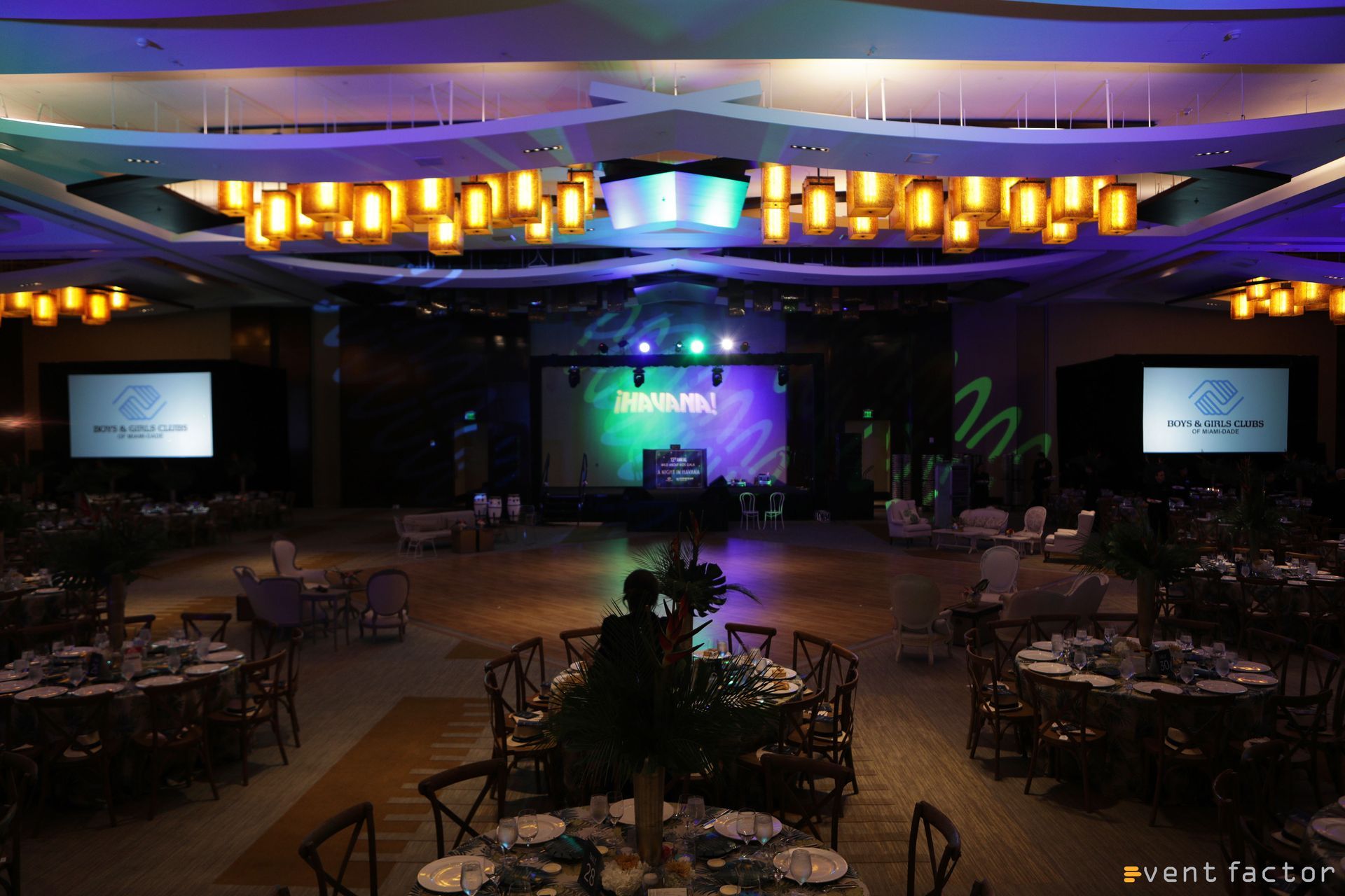 Dimly lit banquet hall with round tables, stage lights, and projection screens at the front