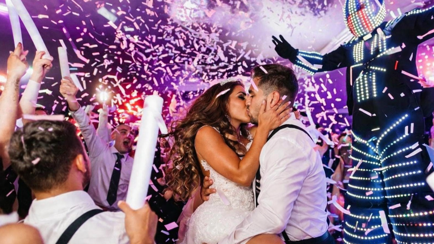 Wedding couple kissing amid confetti as guests cheer and wave glow sticks
