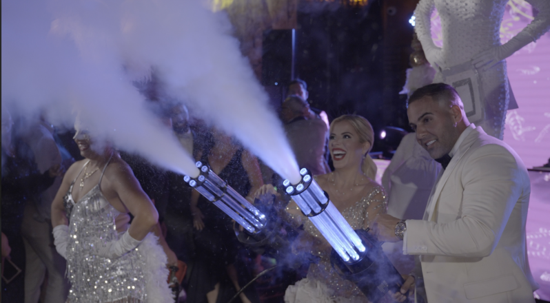 Two performers in black suits fire confetti cannons on a lit stage with band in the background