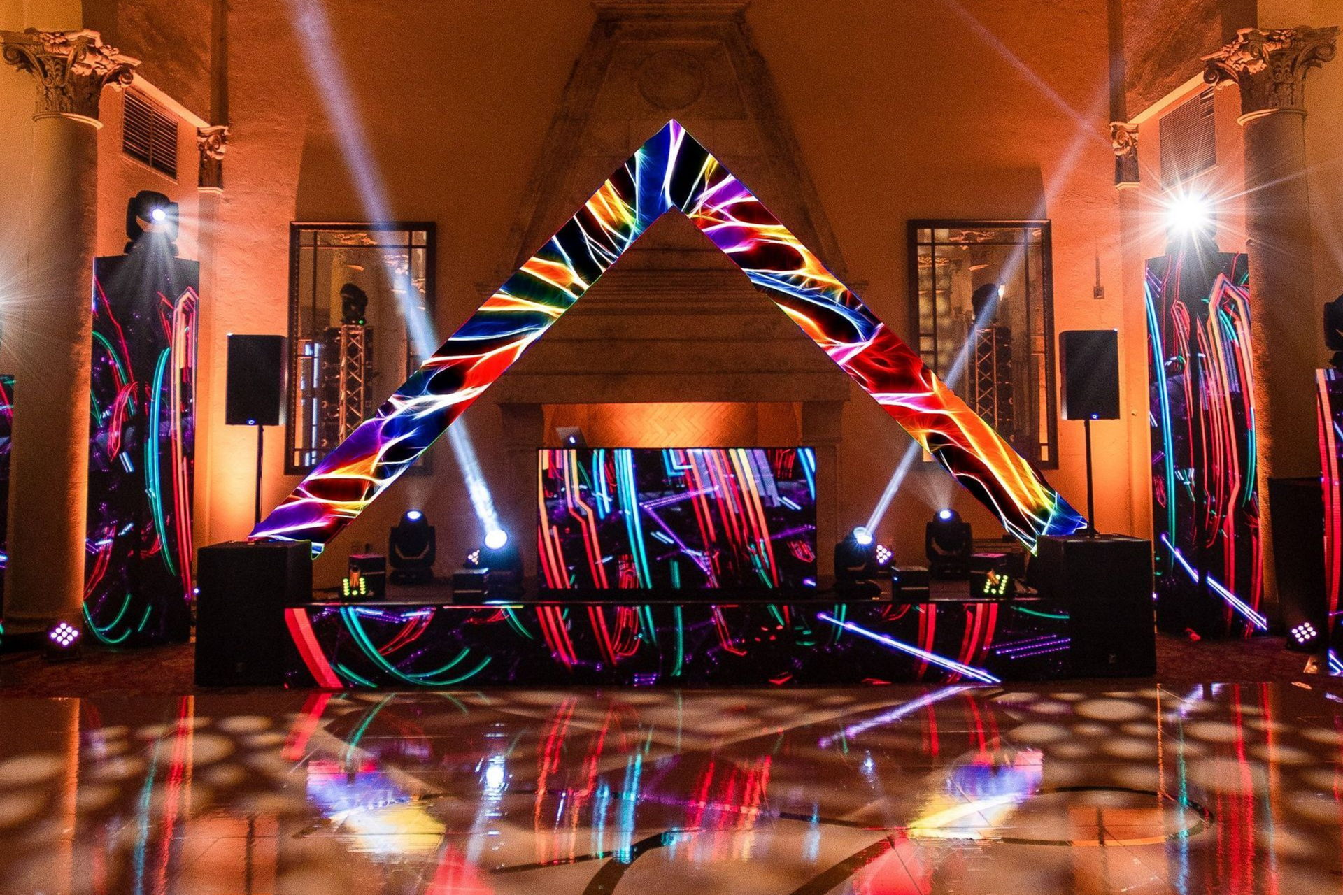 Colorful geometric light installation in a dim indoor hall with a reflective floor and stage setup