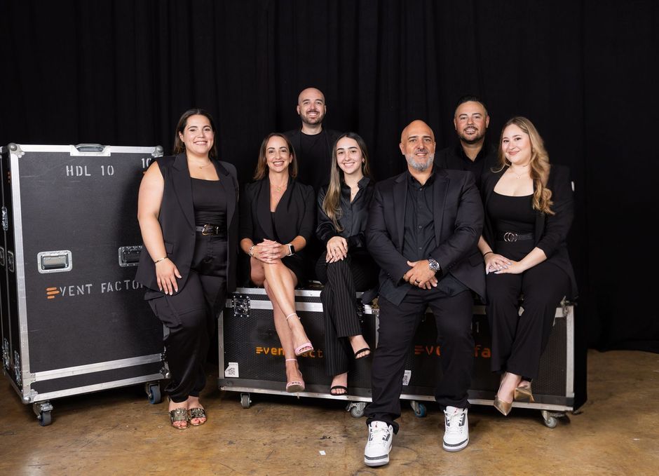 Seven people posing in black outfits on a stage beside black equipment cases, smiling at the camera.