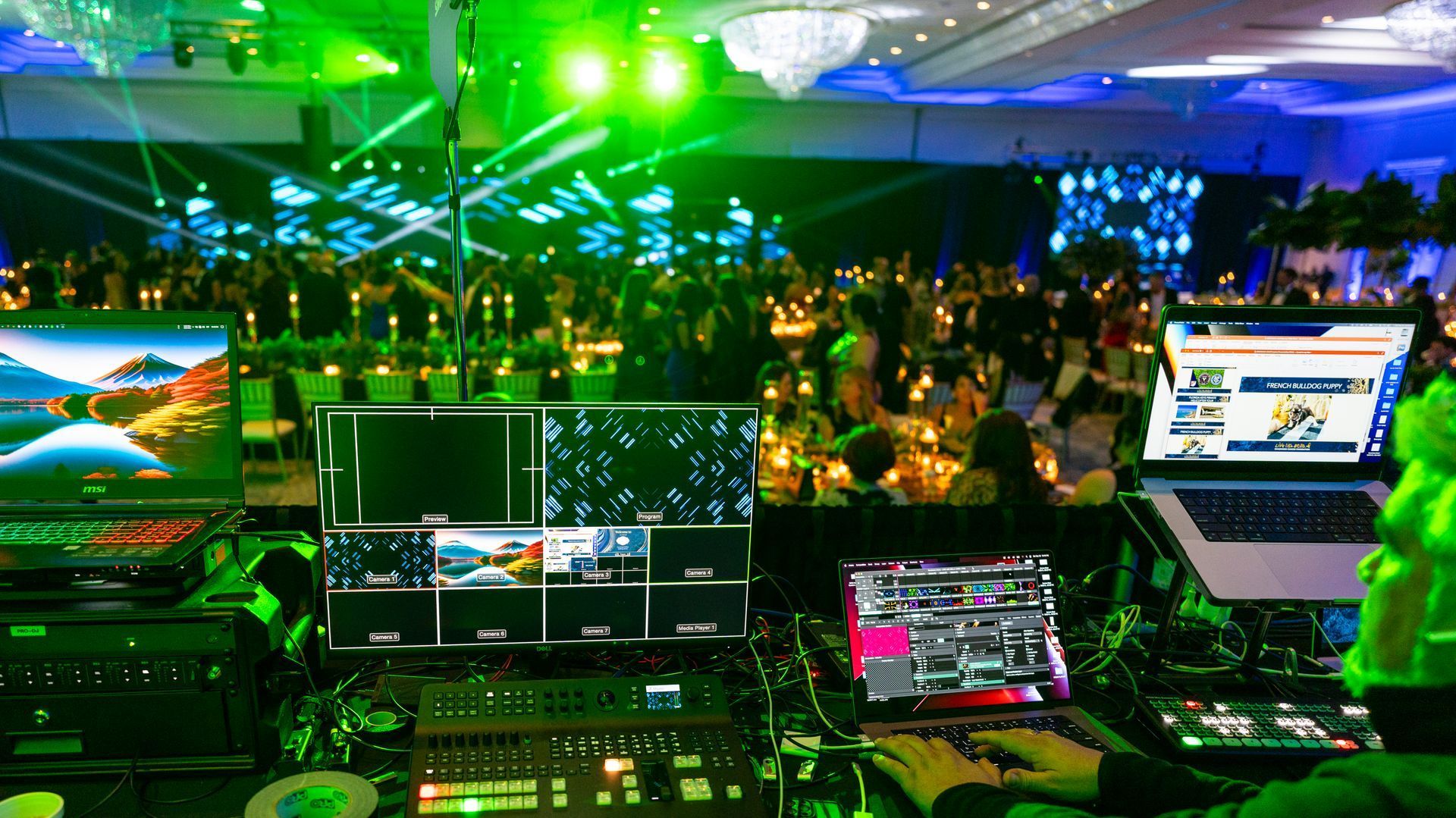 DJ booth overlooking a neon green-lit crowd at a nightclub or concert, with laptops and audio gear in front