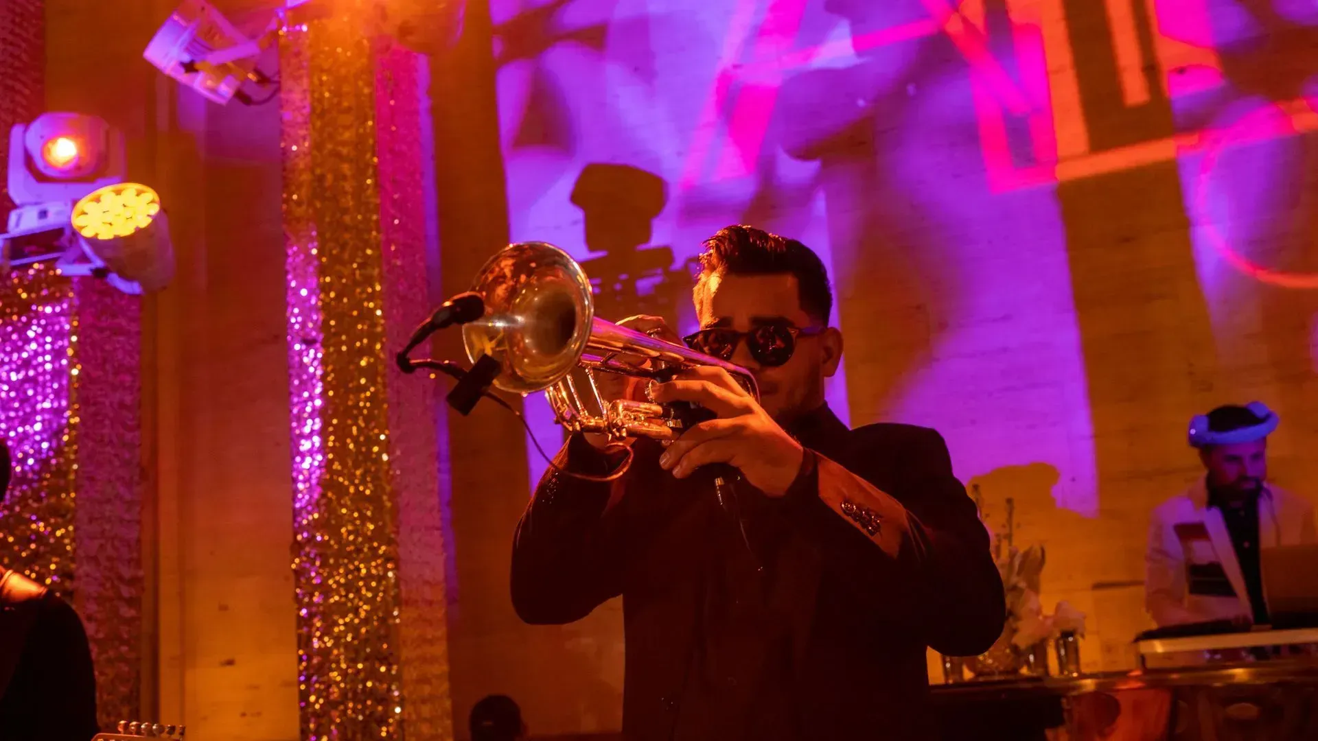 Musician in sunglasses playing trumpet on a colorful stage with warm purple and orange lights