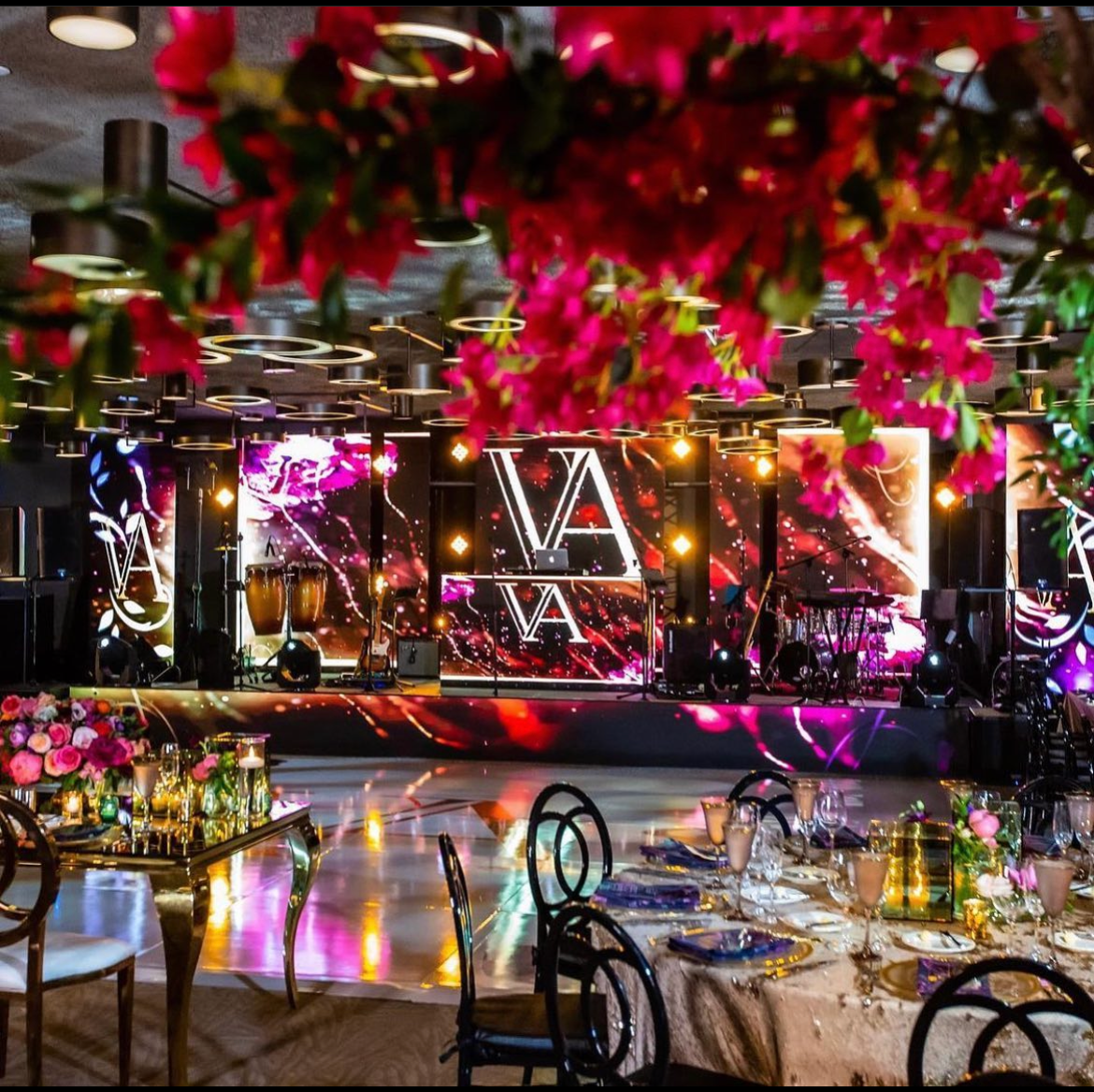 Elegant banquet hall with floral decor, illuminated stage, and set dining tables with glassware and candles
