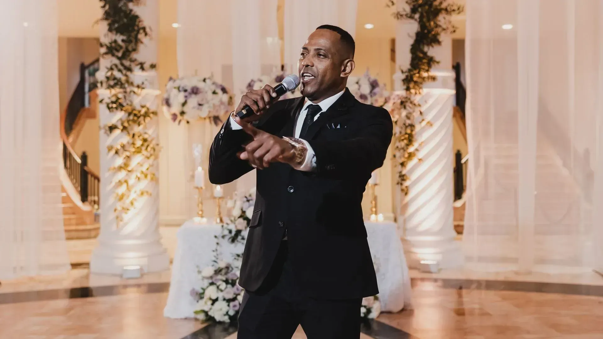 Man in a black suit singing into a microphone at an elegant indoor event with floral decor