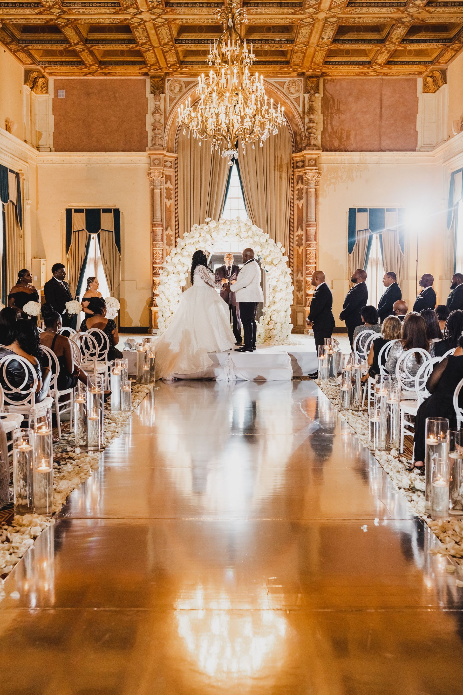 Bride and groom walking down a palm-lined aisle with white floral arrangements and mirrored flooring at sunset