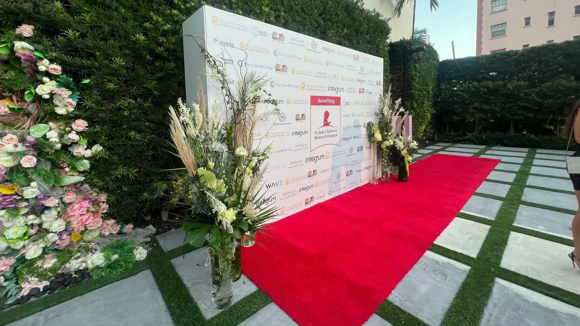 Red carpet walkway beside a floral event backdrop and signage outdoors.