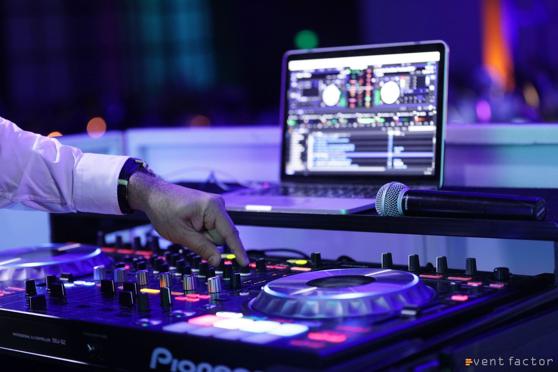 DJ hand adjusting a turntable beside a laptop in a purple-lit club booth