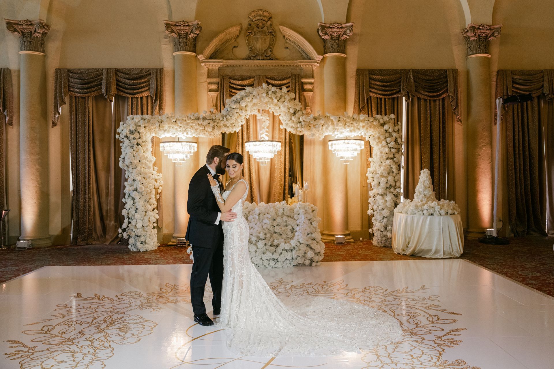 Bride and groom kissing in a gold-lit wedding hall with floral arch and white decor
