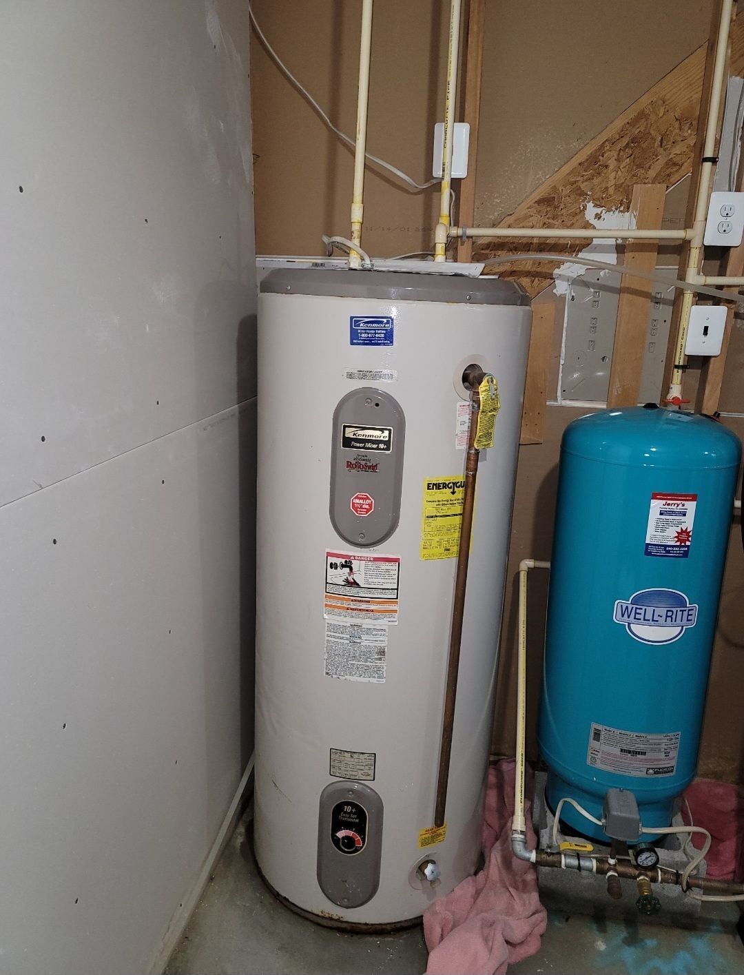 Water heater and blue expansion tank in a utility room. Beige walls, copper pipes.