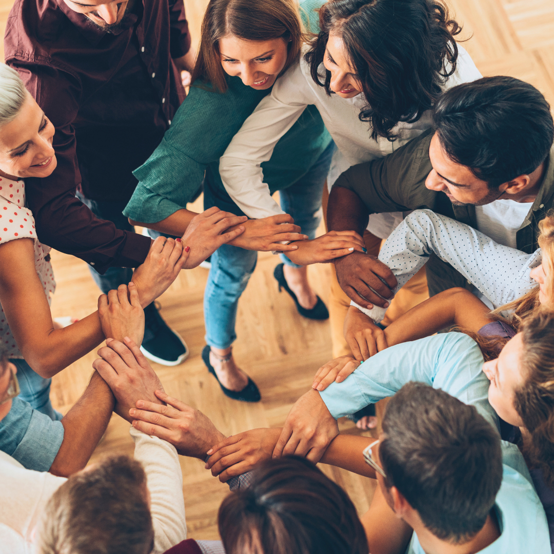 Group of people standing in a circle, hands clasped, heads down, smiling and looking down.