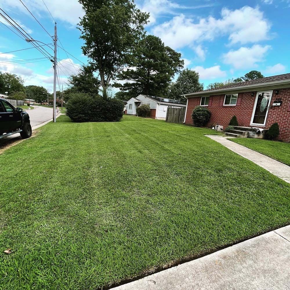 Lush green lawn with freshly mowed stripes, brick house, sidewalk, street, utility pole, and cloudy blue sky.