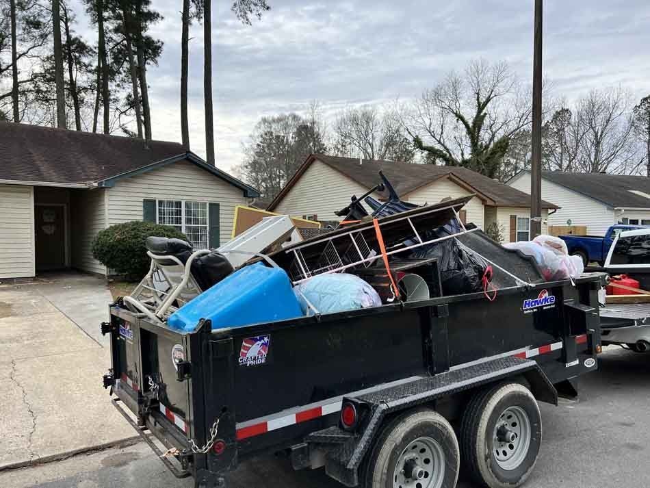 Black trailer filled with discarded household items parked in front of a house. Overcast sky.