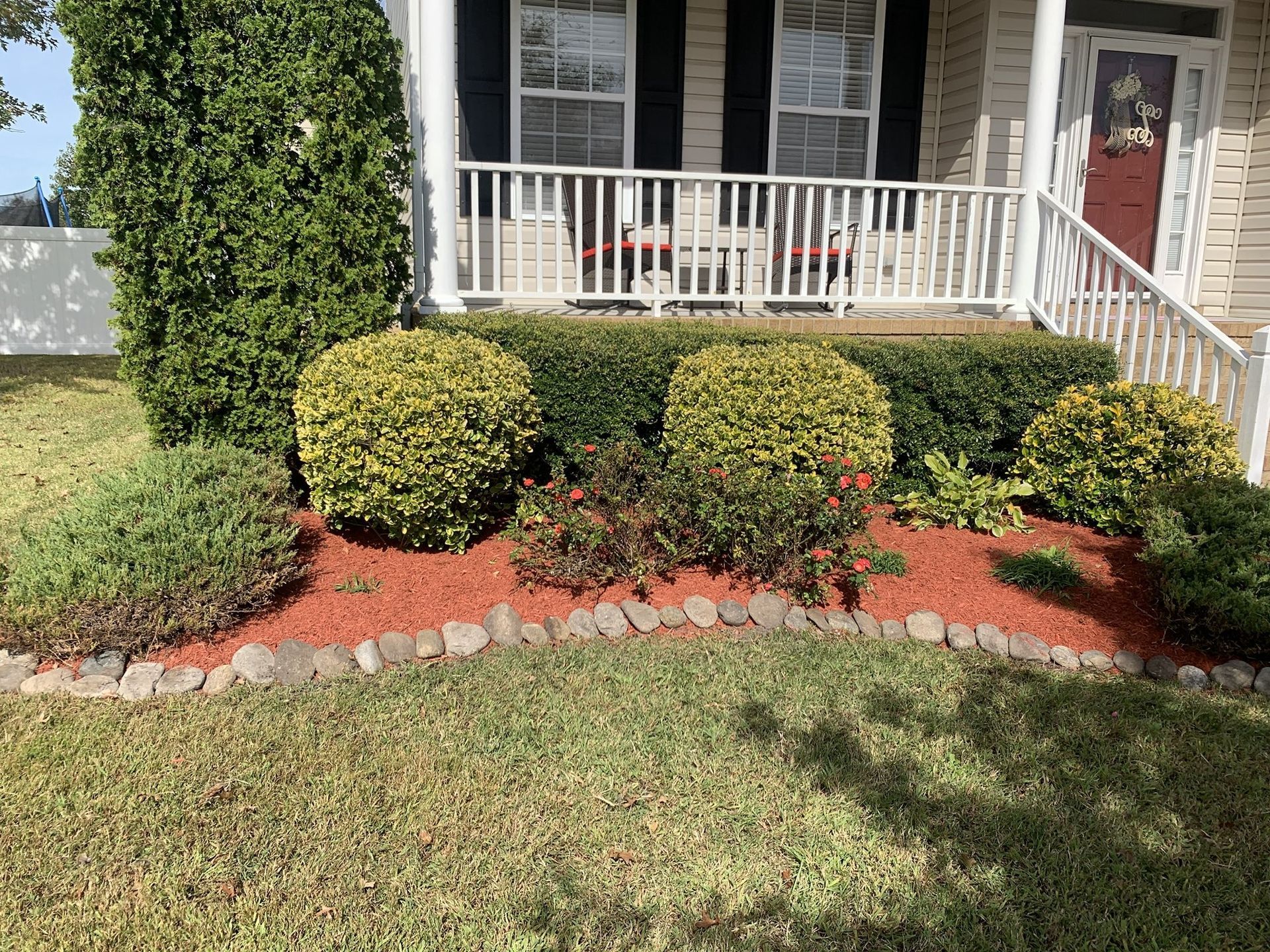 Front yard landscaping with trimmed bushes, red mulch, and stone edging in front of a house.