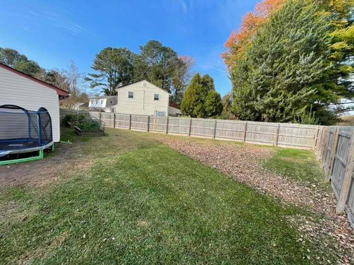 Backyard with green grass, wooden fence, trampoline, and trees under a blue sky.