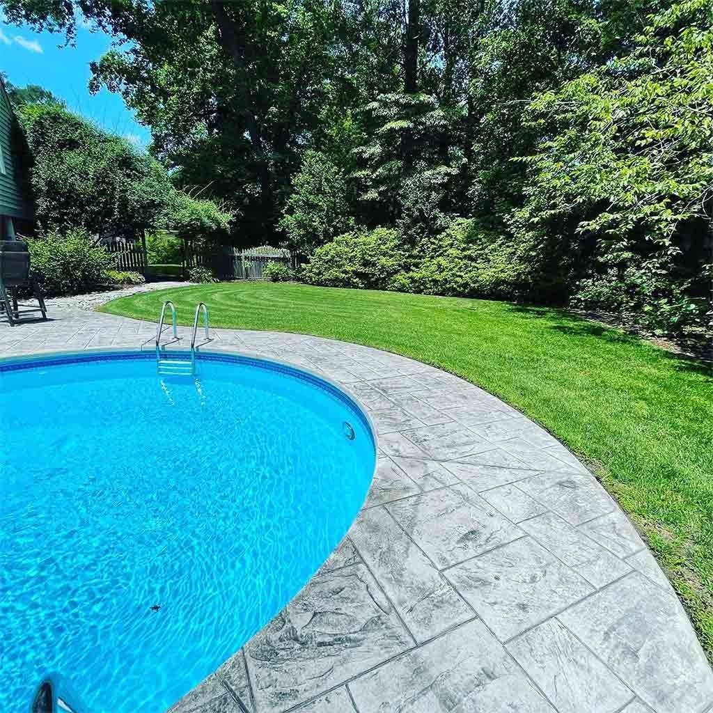 Pool with blue water and stone patio surrounded by green grass and trees.