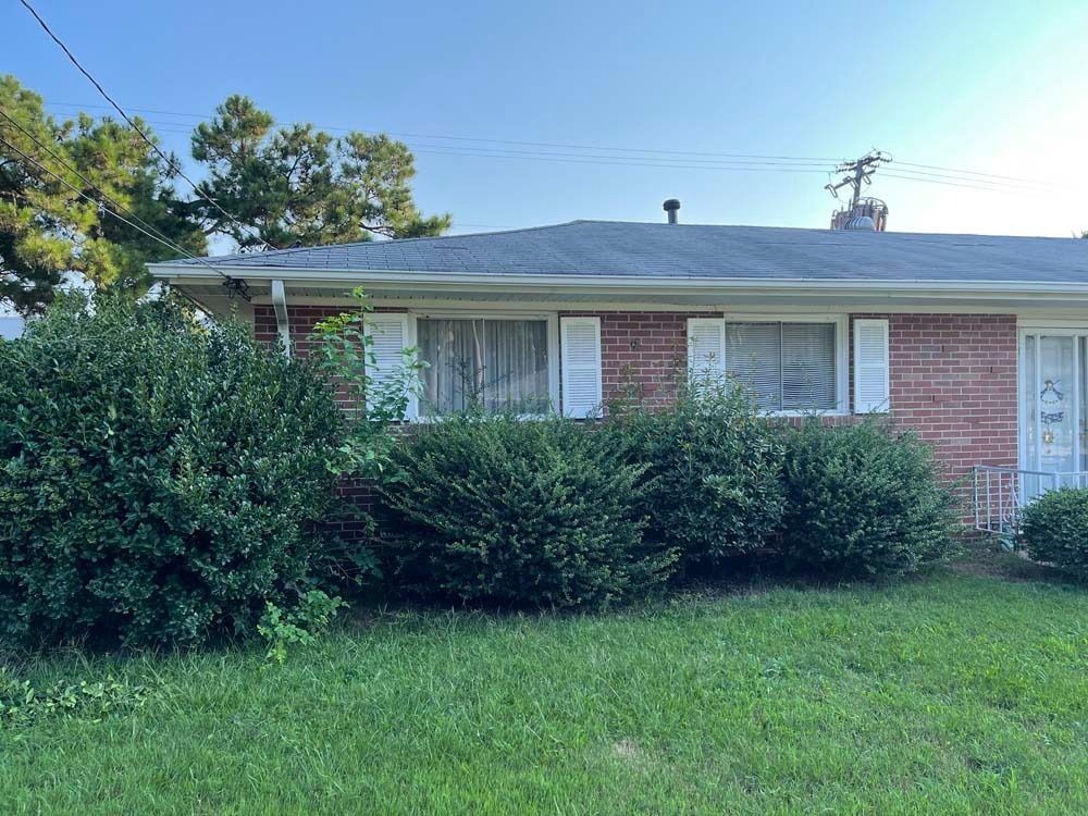 Red brick house with white shutters, overgrown bushes in front, blue sky.