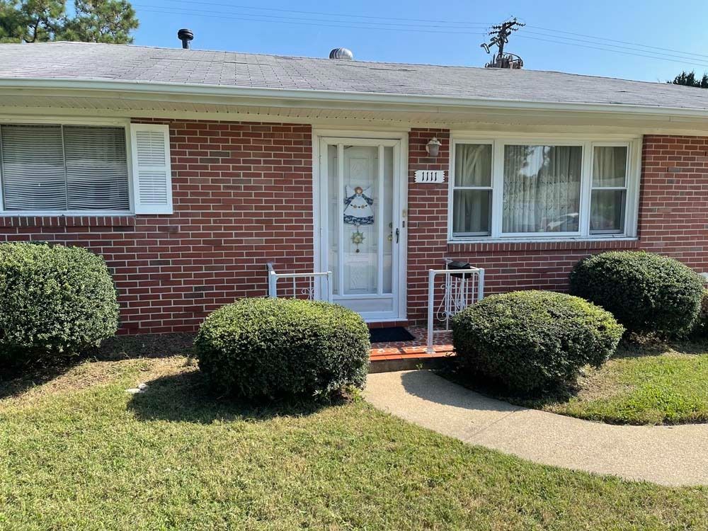 Brick house with white trim, door, and shutters. Three green bushes in front.