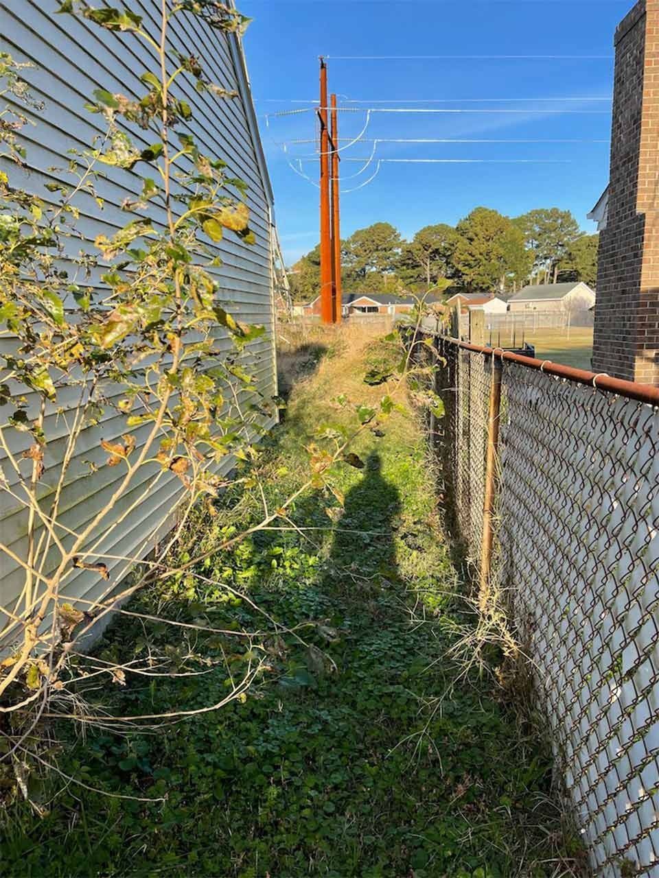 A narrow overgrown alley between a house and a fence, with a power pole in the distance.