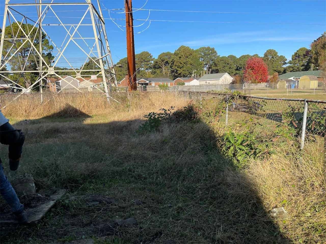 Grassy field next to chain-link fence, power lines, and houses under a clear sky.