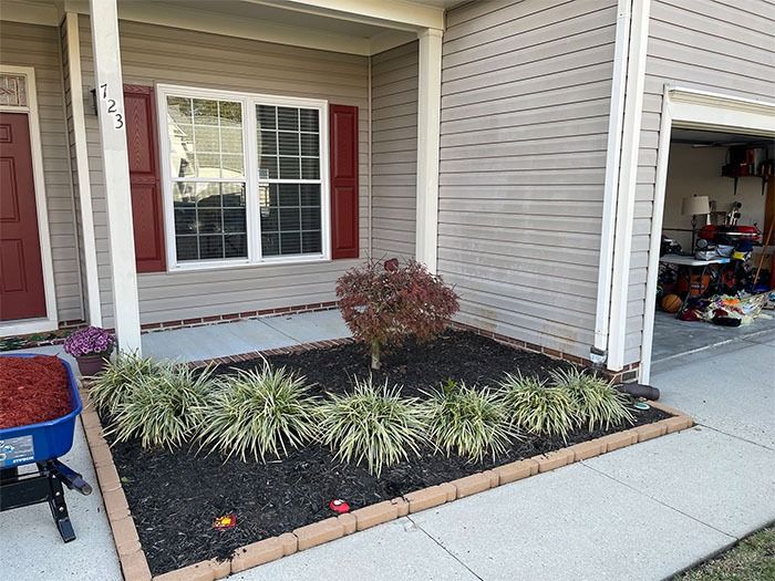 Front yard landscaping: plants with brown mulch bed, brick border, under a house's porch with gray siding.