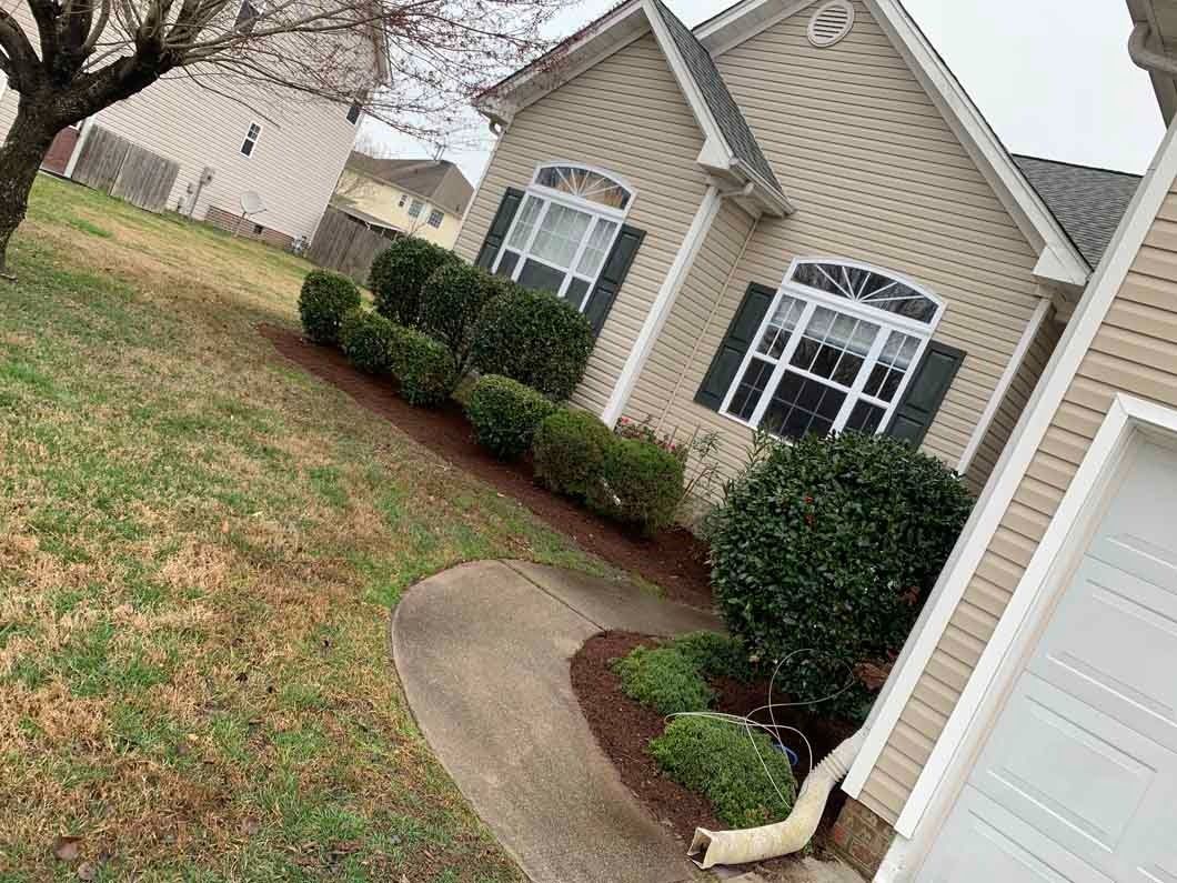 Beige house with green shrubs, dark mulch, and a concrete path.
