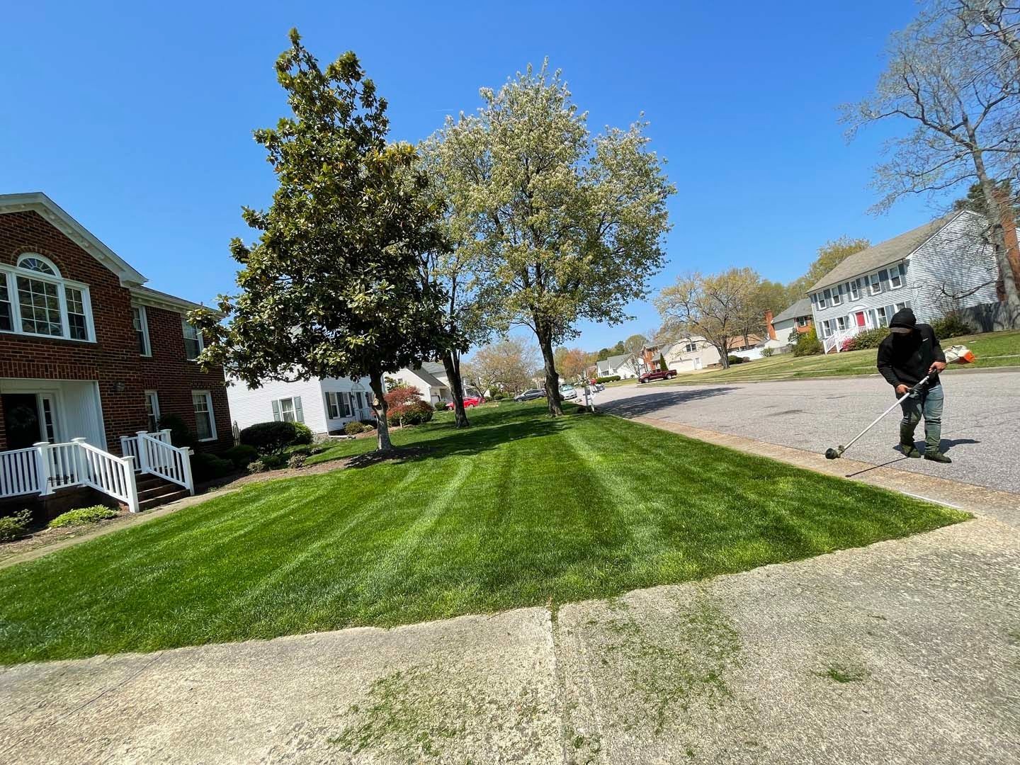 Person mowing a sloped lawn in a residential area; bright green grass, trees, buildings, and blue sky.