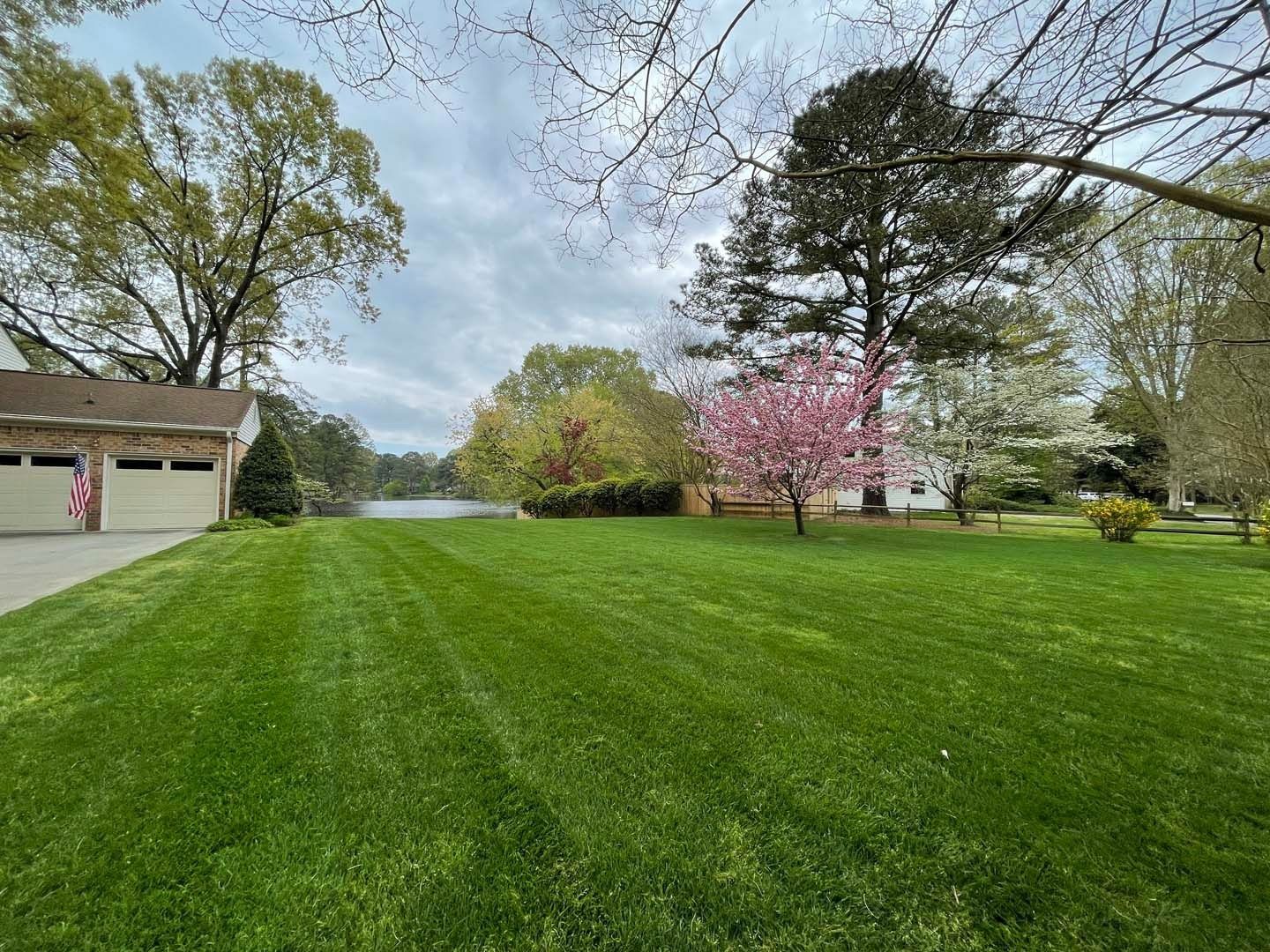 Lush green lawn with a tree in bloom, a house with a garage, and trees under a cloudy sky.
